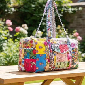 May include: A patchwork tote bag with a rectangular shape and two handles. The bag features a variety of floral and paisley patterns in red, yellow, blue, and green. The handles are striped with black and white. The bag is on a wooden table.