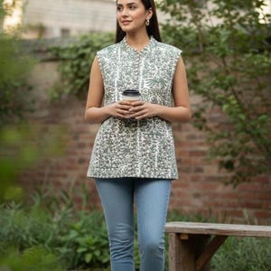 May include: A sleeveless, quilted vest with a white and green floral pattern. The vest is paired with light blue jeans and white sneakers. The person is holding a coffee cup. The background includes greenery, a brick wall, and a wooden bench.