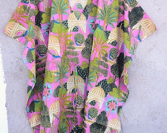 May include: A pink, short-sleeved kaftan with a vibrant print featuring peacocks, trees, and architectural elements. The design incorporates shades of green, beige, and blue, creating a colorful and eye-catching pattern. The kaftan is hanging on a wooden hanger.