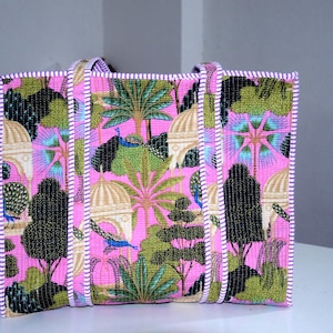 Puede incluir: Bolso tote rosa con un estampado colorido con árboles, edificios y pavos reales. El bolso tiene asas y ribetes a rayas blancas y rosas. El bolso es rectangular y parece estar hecho de tela.