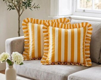 May include: Two square throw pillows with vertical yellow and white stripes and ruffled edges. The pillows are on a gray sofa in a well-lit room. The pillows add a pop of color to the neutral-toned living room.