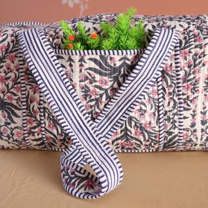 Puede incluir: Bolso tote acolchado con un estampado floral en negro y rosa sobre un fondo blanquecino. El bolso tiene asas a rayas azul marino y blanco. Se ve vegetación y pequeños detalles en naranja dentro del bolso.