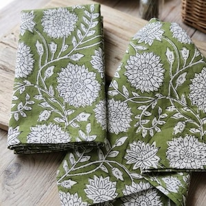 May include: Two folded cloth napkins featuring a repeating pattern of white floral blossoms and leaves printed on a solid olive green background. The fabric displays a block-print style design with visible white outlines on the petals and foliage.