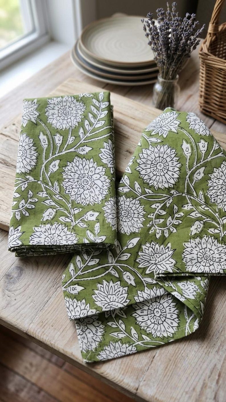 May include: Two folded cloth napkins featuring a repeating pattern of white floral blossoms and leaves printed on a solid olive green background. The fabric displays a block-print style design with visible white outlines on the petals and foliage.