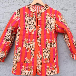 以下が含まれることがあります： 鮮やかなオレンジとピンクのキルティングジャケット、トラ柄。 ジャケットはマンダリンカラー、長袖、フロントポケット2つ。 トラ柄は黒とタンで、木製のハンガーに掛けられています。