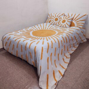 May include: A white and light blue bedspread and pillow set with a large sunburst design in yellow. The bedding set includes a comforter and two matching pillows, creating a cohesive and decorative bedroom ensemble.