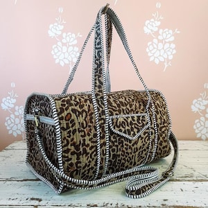 May include: A quilted duffel bag with a leopard print pattern in brown and tan. The bag has black and white striped trim, handles, and a detachable shoulder strap. The bag is on a white wooden surface.