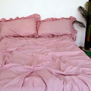 May include: A pink ruffled bedspread with two matching ruffled pillows. The bedspread is made of a soft, smooth fabric and has a slightly wrinkled appearance.