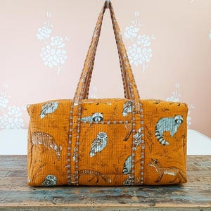 Könnte beinhalten: Senffarbene Tragetasche mit einem Waldtiermuster. Die Tasche zeigt Illustrationen von Hirschen, Eulen, Waschbären und Füchsen. Die Tasche hat zwei Griffe und eine Tasche auf der Vorderseite. Die Tasche ist aus einem gesteppten Stoff gefertigt.
