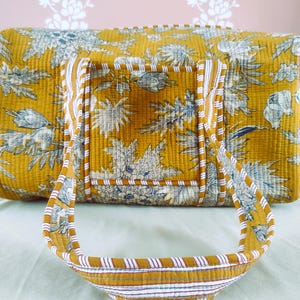 Puede incluir: Bolso de lona acolchado amarillo mostaza con un estampado floral en blanco y azul. El bolso tiene asas a juego con rayas blancas y rojas. El bolso está hecho de una tela suave y acolchada.