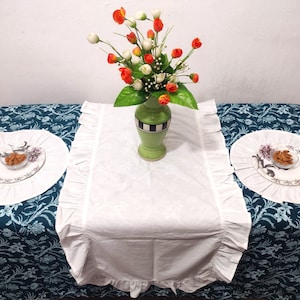 以下が含まれることがあります： フリル付きの白いテーブルランナーが、花柄のターコイズブルーのテーブルクロスの上に置かれています。ランナーの中央には、オレンジと白の花が活けられた花瓶が置かれています。ランナーの両側には、アーモンドの入った小さなボウルが置かれた白い丸いプレースマットが2つあります。