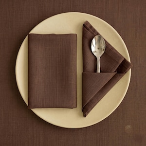 May include: A round, cream-coloured plate holds two dark brown fabric napkins with a visible woven texture. One napkin is folded into a rectangle, while the other is folded into a triangle with a silver-toned metal spoon resting on top.