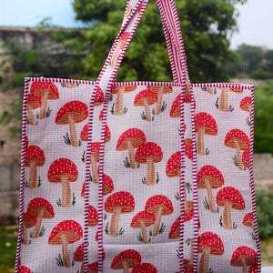 Puede incluir: Un bolso tote blanco con un patrón repetido de setas rojas. El bolso tiene asas y ribetes a rayas rojas y blancas. El fondo es una escena borrosa al aire libre.