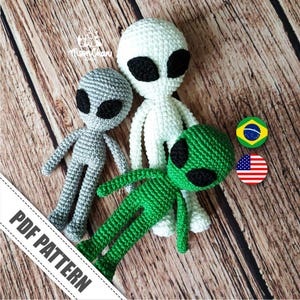 May include: Three crocheted alien amigurumi toys. One is white, one is gray, and one is green. The toys are sitting on a wooden surface.  PDF PATTERN.