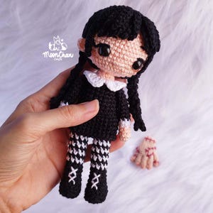 May include: A crocheted doll with black hair in plaits, dressed in a black dress with a white collar and patterned leggings. The doll has a light skin tone and large black eyes. A detached crochet hand is also visible. The doll is held in a hand.