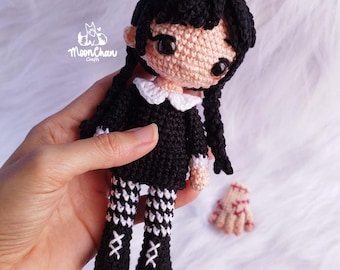May include: A handmade crochet doll with black hair in braids, wearing a black dress with a white collar and patterned leggings. The doll has a light skin tone and large black eyes. A detached crochet hand is also visible. The doll is held in a hand.