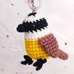 May include: A handmade crochet bird keychain in yellow, black, white, and brown. The bird has a black beak, black eyes, and black feet. The keychain is attached to a clear plastic ring. The bird's body is yellow, with a white and black head.