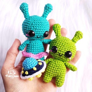 Op de afbeelding: Twee gebreide alien knuffels, een groene en een blauwe, met grote zwarte ogen en glimlachende gezichten. De blauwe alien houdt een gebreide vliegende schotel vast.