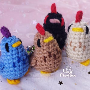 Puede incluir: Cuatro llaveros de pollo de ganchillo hechos a mano en azul, marrón, negro y blanco. Cada pollo tiene una cresta roja, un pico amarillo y ojos negros. Los llaveros están sobre una superficie blanca con un logo "MoonChan crafts".