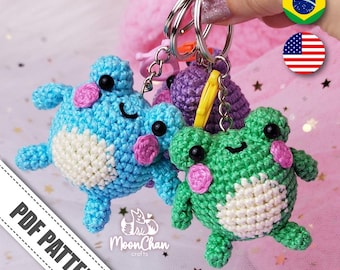 May include: Three crocheted frog keychains in blue, green, and purple. The keychains are attached to a silver key ring. The keychains are on a pink background with white stars.