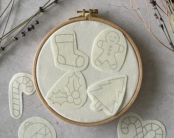 May include: A wooden embroidery hoop with a cream-colored fabric, featuring several Christmas-themed felt cutouts. These include a stocking, gingerbread man, candy cane, Christmas tree, ornaments, and a bell with holly.