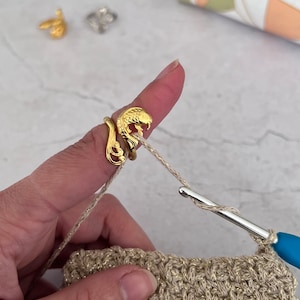 Op de afbeelding: Een gouden ring met een visontwerp wordt gebruikt om garen vast te houden tijdens het haken van een beige gehaakt stof.