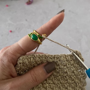 Könnte beinhalten: Nahaufnahme einer Hand, die ein Häkelprojekt hält. Die Person trägt einen goldenen Ring mit einem grünen Stein. Das Häkelprojekt ist aus beigefarbenem Garn gefertigt und hat ein strukturiertes Muster.