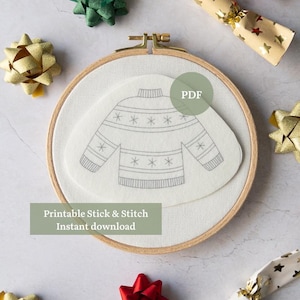 Peut inclure: Un cercle à broder en bois avec un tissu blanc et un motif de pull gris. Le texte "Printable Stick & Stitch Instant download" est visible. Des nœuds dorés, verts et rouges et des crackers à motifs d'étoiles entourent le cercle.