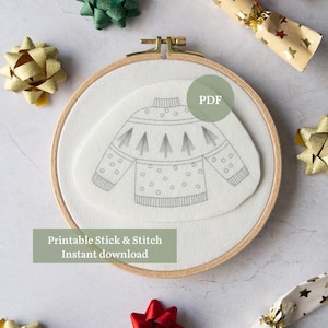 Peut inclure: Un cercle à broder en bois avec un motif de pull gris sur tissu blanc. Le texte "Printable Stick & Stitch Instant download" est affiché. Des nœuds et des pétards décoratifs sont disposés autour du cercle.