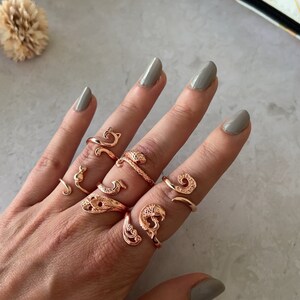 Könnte beinhalten: Eine Hand trägt fünf goldfarbene Ringe mit verschiedenen Designs, darunter eine Katze, eine Schlange und ein Wirbel. Die Ringe werden am Mittel- und Ringfinger getragen.