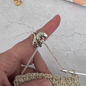 Puede incluir: Un anillo porta-ganchillo de plata con forma de pez en un dedo que sostiene un hilo beige. El hilo está unido a una tela de crochet beige.