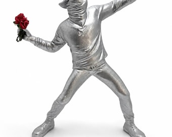 May include: A silver-colored sculpture of a figure in a baseball cap and face covering, holding a bouquet of red roses in one hand and extending the other. The figure is wearing a long-sleeved shirt, pants, and boots.