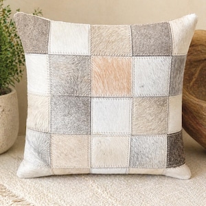May include: Square decorative pillow with a patchwork design. The pillow features squares of various colors, including white, gray, beige, and brown. The squares are stitched together with visible seams. The pillow has a neutral color palette.