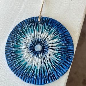 Puede incluir: Adorno de pared redondo y texturizado con un diseño radial en tonos azules, blancos y turquesas. El centro presenta un pequeño círculo azul claro. Tiene un lazo de cuerda natural para colgar. Sobre una superficie blanca.