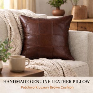 May include: A brown, square leather pillow with a patchwork design rests on a beige sofa. The pillow is made of genuine leather and has a grid-like pattern. The text at the bottom reads "HANDMADE GENUINE LEATHER PILLOW" and "Patchwork Luxury Brown Cushion."