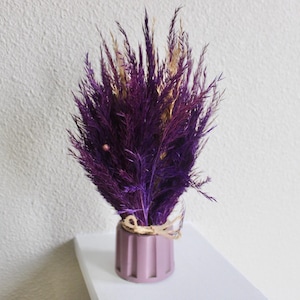 May include: A decorative arrangement of dried purple and gold-toned grasses in a small, ribbed, mauve-colored vase. The vase is tied with twine and sits on a white shelf against a textured white wall.