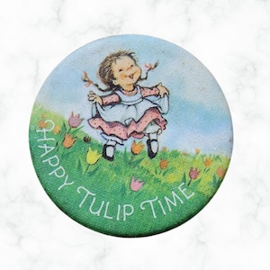Puede incluir: Botón redondo con una ilustración vintage de una niña con un vestido blanco y rosa, saltando en un campo de tulipanes. El texto "Happy Tulip Time" está impreso alrededor del borde.