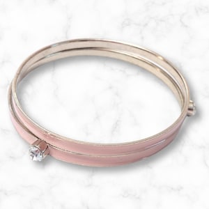 Può includere: Un braccialetto a cerchio rosa chiaro con una fascia in metallo argentato. Il braccialetto presenta un singolo accento di cristallo trasparente a taglio quadrato. Il braccialetto è un gioiello semplice ed elegante.