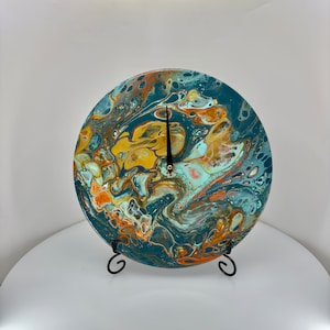 Könnte beinhalten: Runde Wanduhr mit einem einzigartigen abstrakten Design. Das Zifferblatt zeigt ein wirbelndes Muster aus Türkis, Gold, Orange und Weiß. Schwarze Zeiger zeigen die Uhrzeit an. Die Uhr wird auf einem schwarzen Metallständer präsentiert.