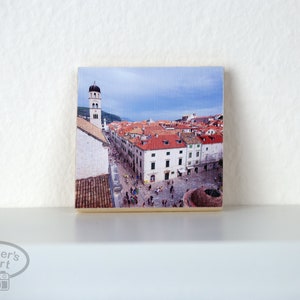 May include: A photograph of a city with red tiled roofs and a tall white tower. The city is surrounded by a wall and there are people walking in the streets.