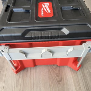 May include: Two stacked Milwaukee Packout storage containers. The top container is black with a red Milwaukee logo. The bottom container is red with grey and silver accents. Designed for tool storage, they are robust and practical.