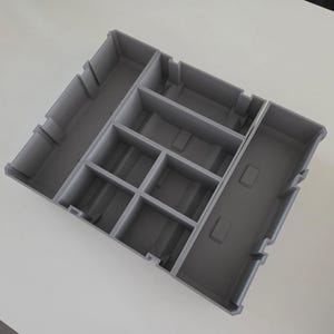 以下が含まれることがあります： さまざまなサイズのコンパートメントが複数あるグレーのプラスチック製オーガナイザー。オーガナイザーは長方形で、一連の仕切りがあり、別々の収納スペースを作成しています。デザインは3Dプリントのようで、マット仕上げです。