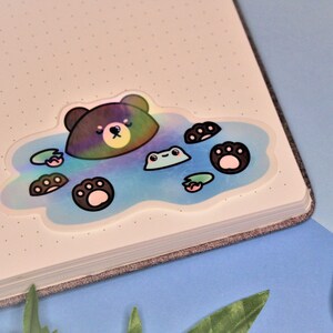 May include: A holographic sticker featuring a brown bear with a blue and white background. The bear is submerged in water with its paws and a frog visible. The sticker is ideal for adding a touch of whimsy to your planner, journal, or laptop.