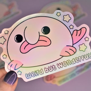 May include: A holographic sticker featuring a cartoon axolotl with a pink tongue and a pink fin. The axolotl is surrounded by stars and the text "Weird but wonderful!" is printed below it.
