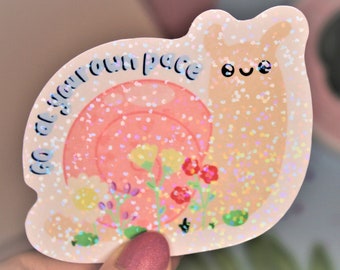 May include: A pink and white iridescent glitter snail sticker with the text "Go at your own pace" in blue. The snail has a cute cartoon face with two eyes and a smile.