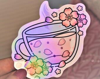 May include: A holographic sticker of a teacup with pink cherry blossoms and a pink and purple iridescent liquid inside. The teacup has a white rim and a black outline.