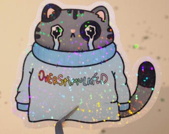 May include: A holographic sticker of a gray cat wearing a blue sweater with the text "OVERSTIMULATED" in rainbow colors. The cat has a sad expression and is crying.