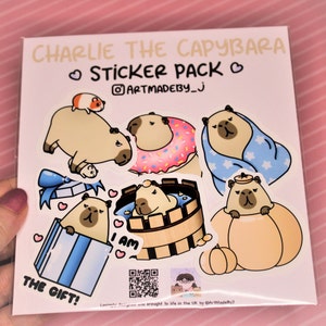 Op de afbeelding: Een stickerpakket met een cartoon capybara in verschillende poses, waaronder met een donut, een badjas en een pompoen. De tekst "Charlie the Capybara Sticker Pack" staat bovenaan het vel. De tekst "I am" en "The Gift!" zijn ook inbegrepen.