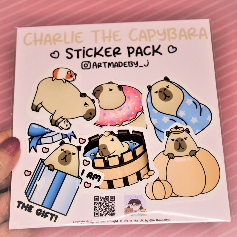 Op de afbeelding: Een stickerpakket met een cartoon capybara in verschillende poses, waaronder met een donut, een badjas en een pompoen. De tekst "Charlie the Capybara Sticker Pack" staat bovenaan het vel. De tekst "I am" en "The Gift!" zijn ook inbegrepen.