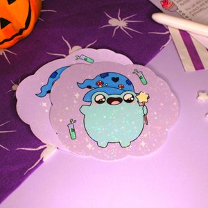 Peut inclure: Deux autocollants holographiques repr&eacute;sentant une grenouille de dessin anim&eacute; portant un chapeau de sorcier bleu avec une &eacute;toile au sommet. La grenouille tient une baguette et est entour&eacute;e d'&eacute;toiles et de paillettes. Les autocollants sont en forme de nuages et sont sur un fond violet.